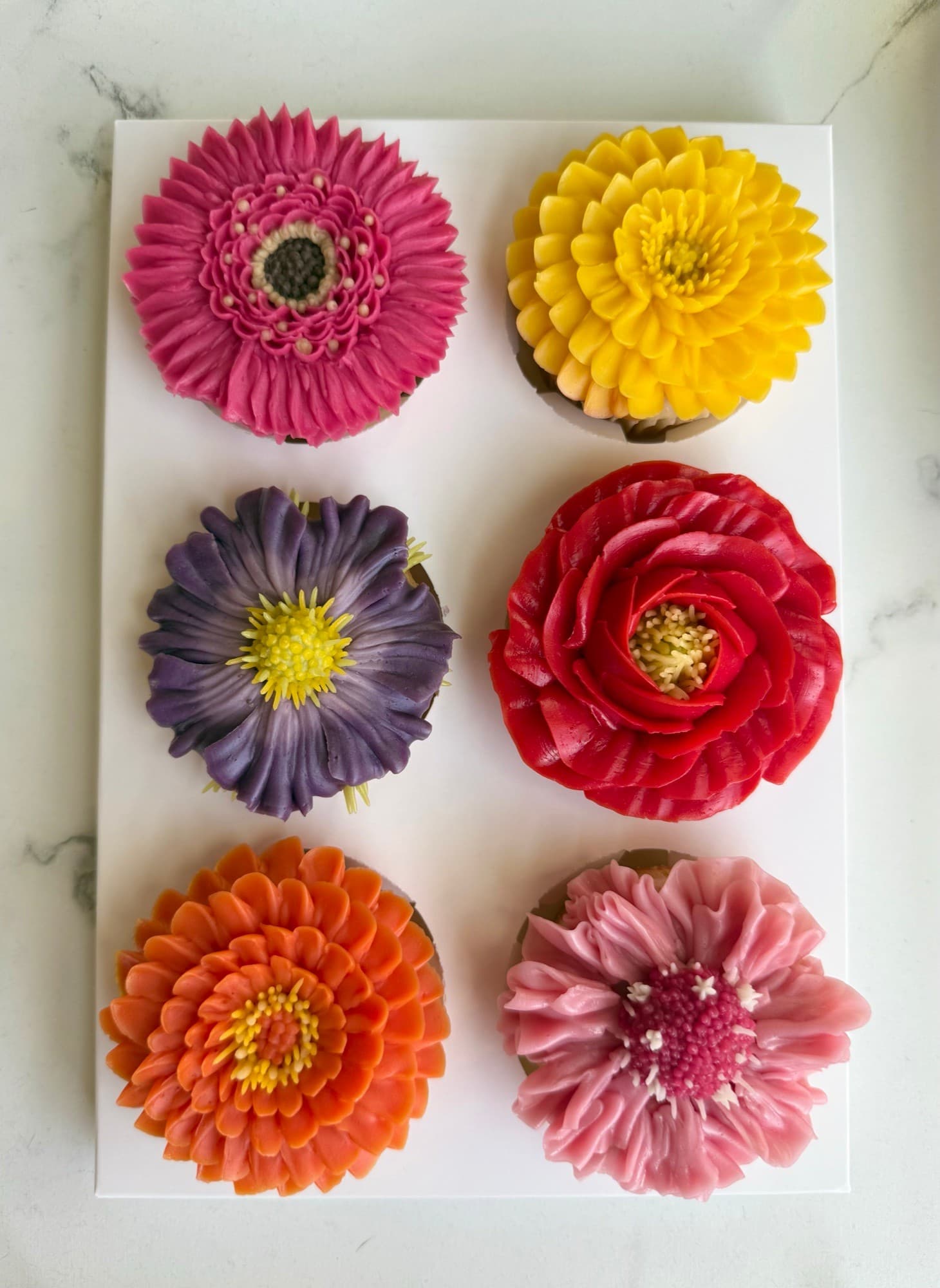 Six vibrant buttercream flower cupcakes in pink, yellow, purple, red and orange