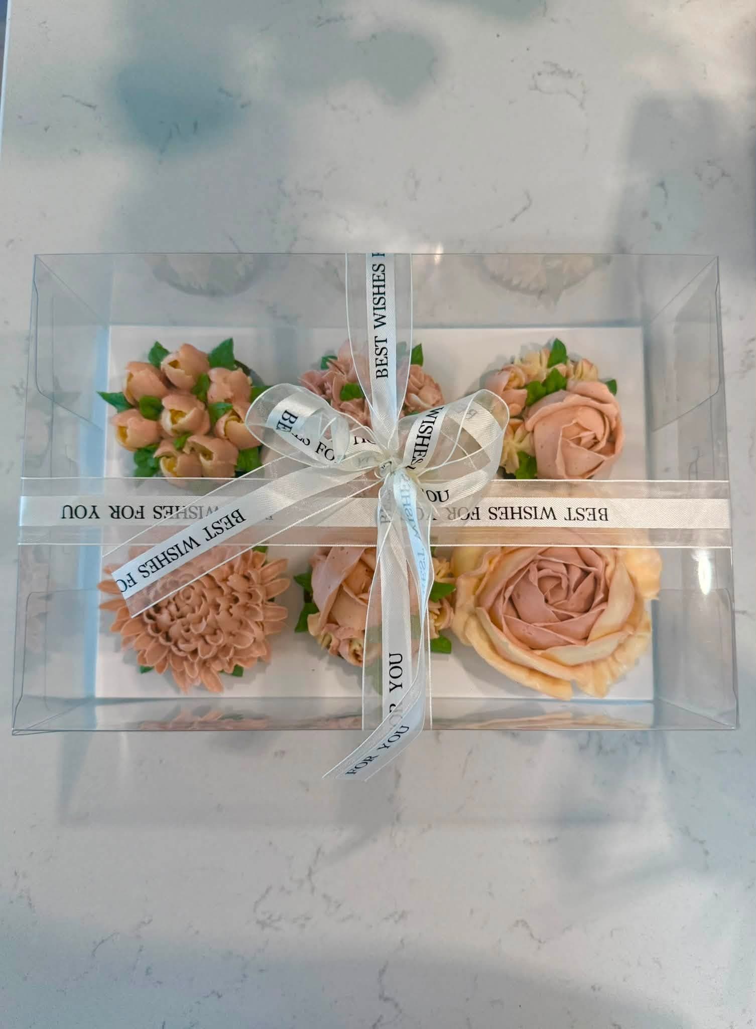 A clear Best Wishes gift box of apricot and cream floral cupcakes