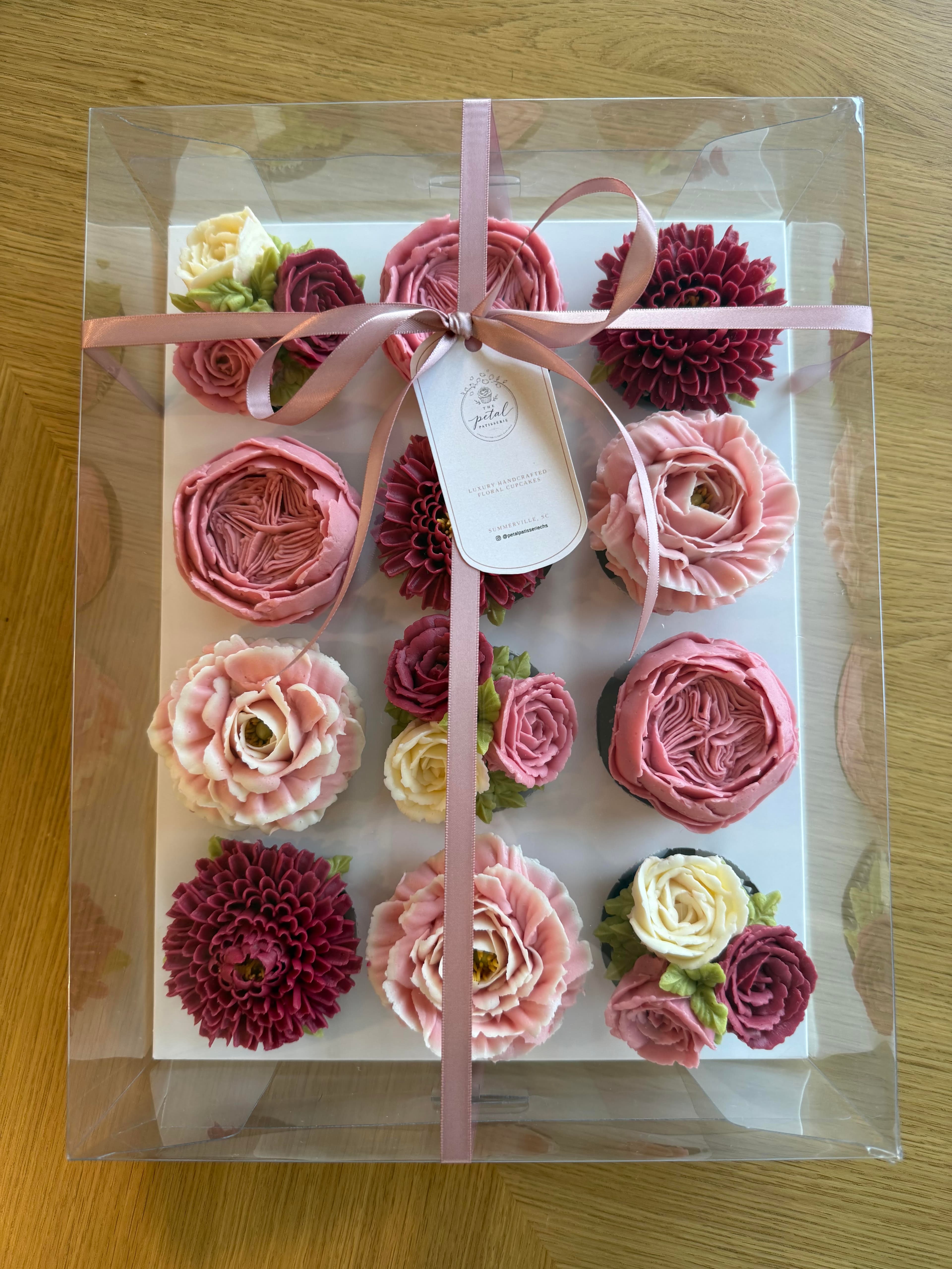 A dozen floral cupcakes in pinks and purples in their gift box