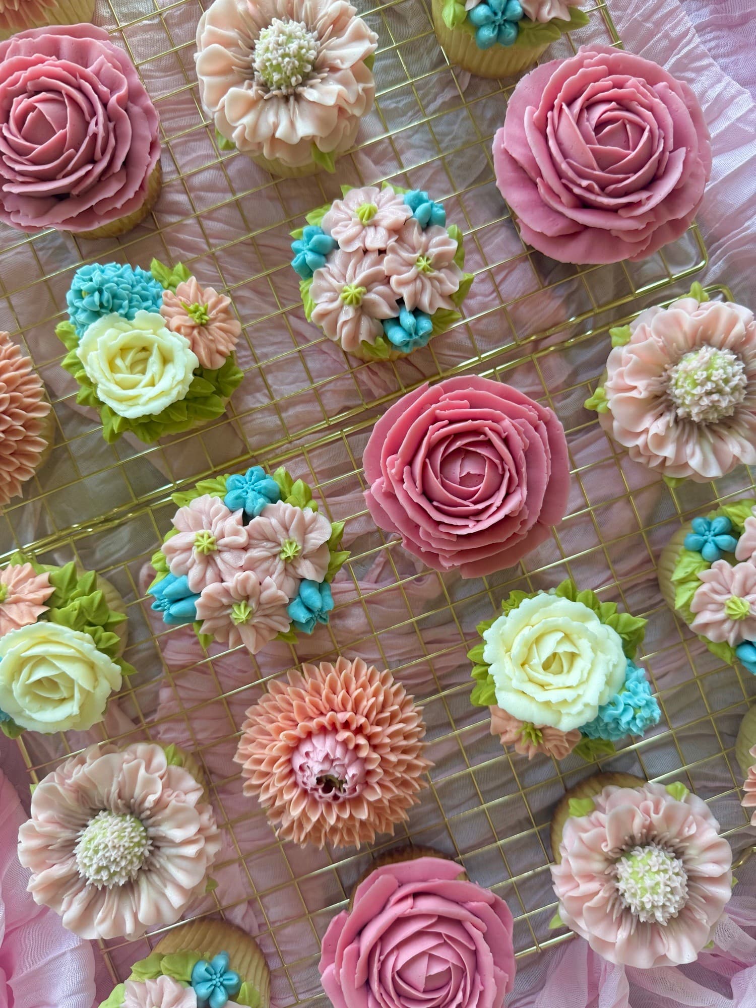 Close-up of pastel buttercream roses and mixed flower cupcakes on gold cooling racks